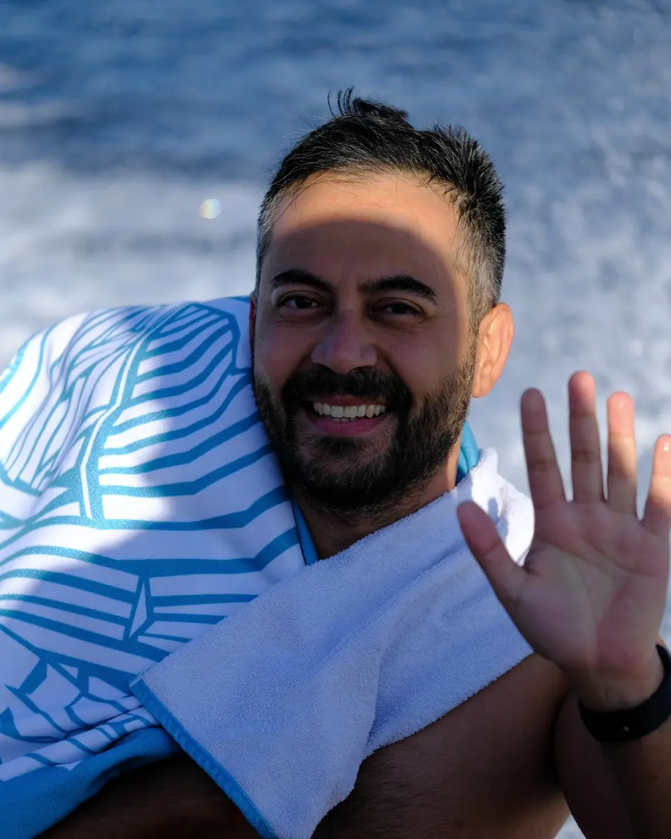 Kolunda havlu olan bir katılımcı gülümseyerek el sallıyor; arkada dalgalar.