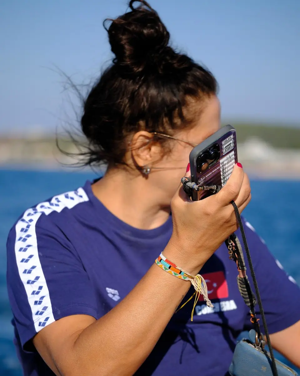 Mavi antrenman tişörtlü biri, telsizi ağzına tutarak konuşuyor; arkada deniz.