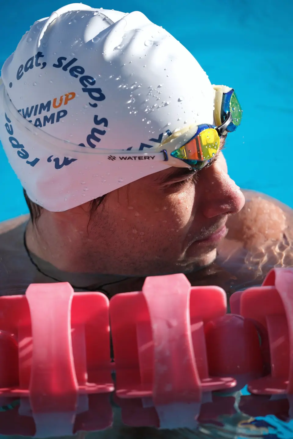 Swim Up Camp baskılı bone takmış yüzücünün yakın çekimi, kulvar şamandıraları önde.