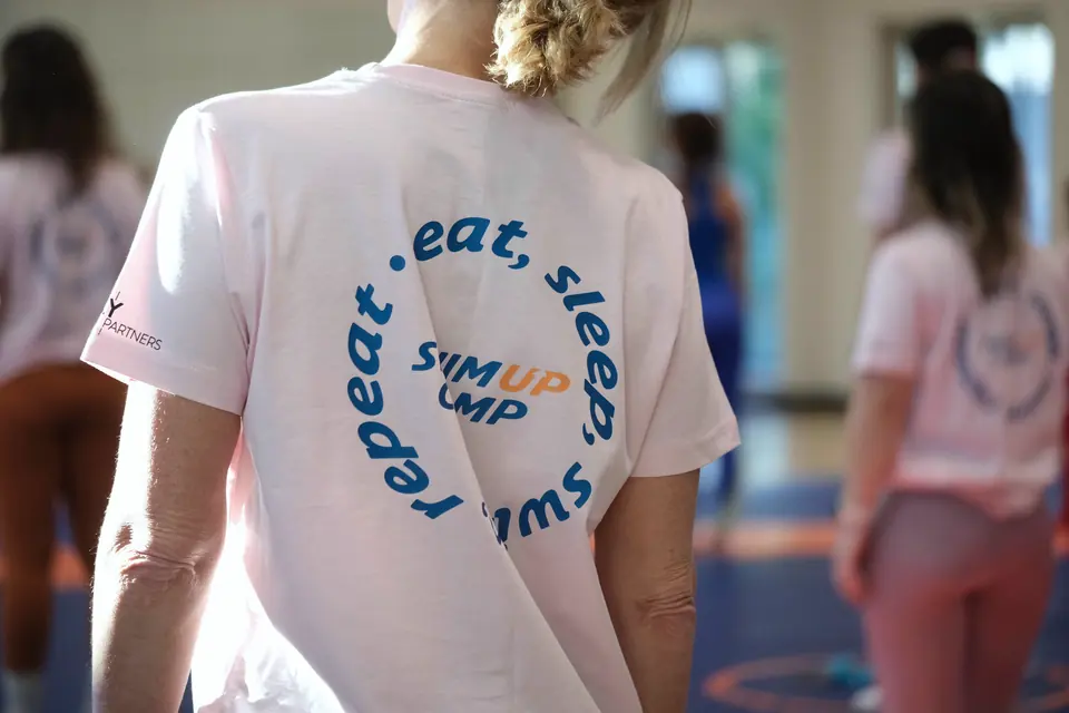Pembe tişörtün sırtında "eat, sleep, swim, repeat" yazısı; salonda katılımcılar.