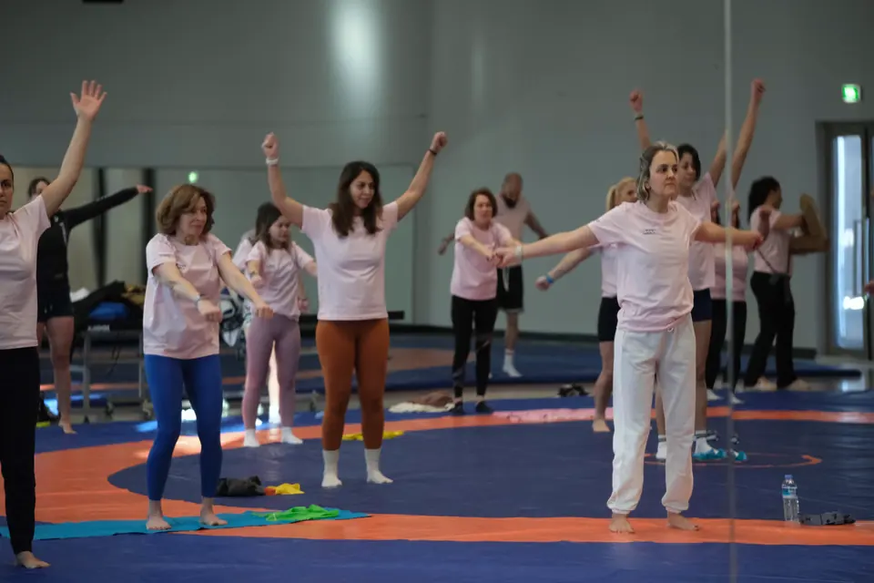 Spor salonunda minderde pembe tişörtlü grup, kollarını açarak ısınıyor.
