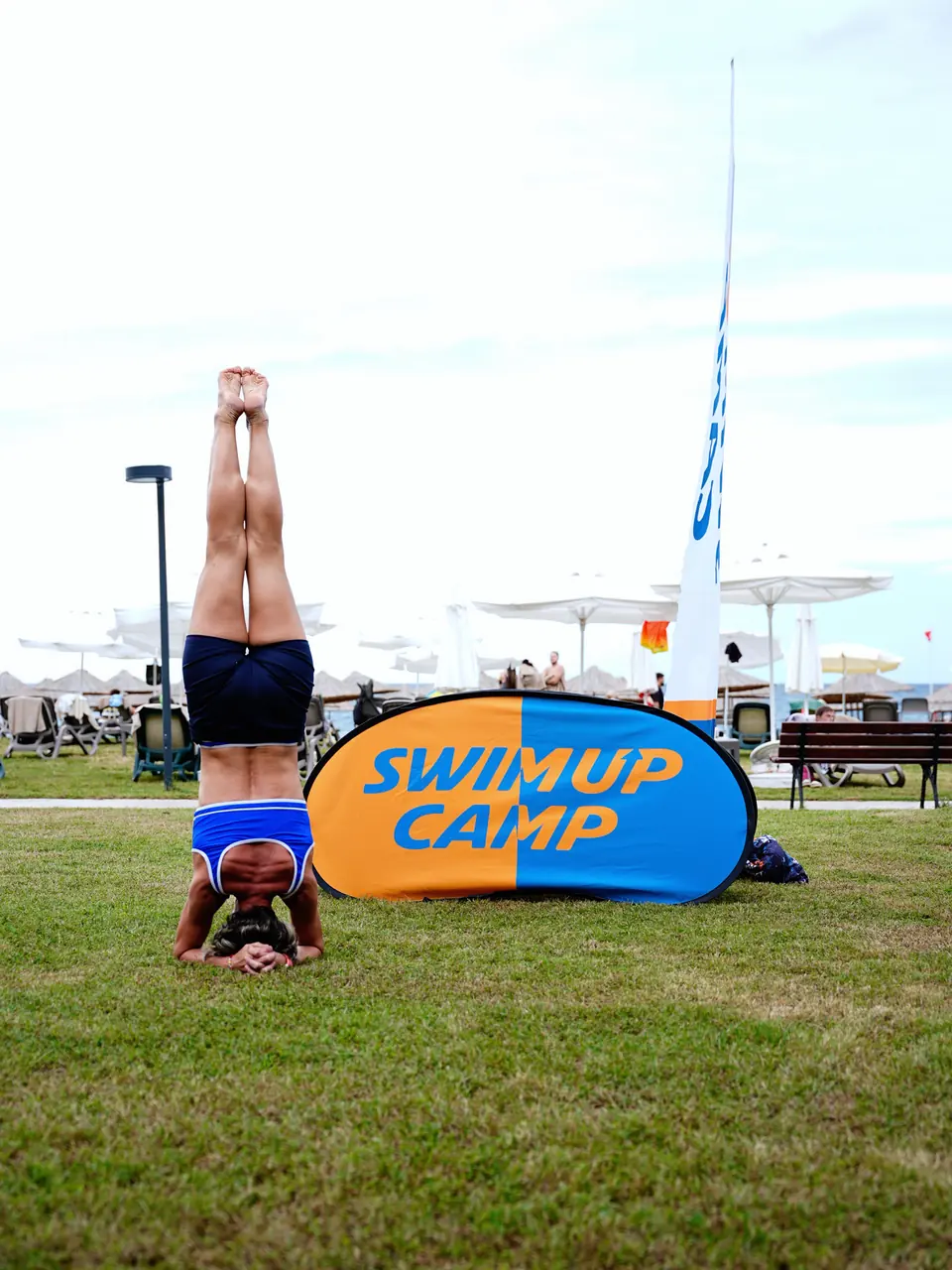 Çimde baş üstü duruşu yapan bir katılımcı, arkada Swim Up Camp tabelası.