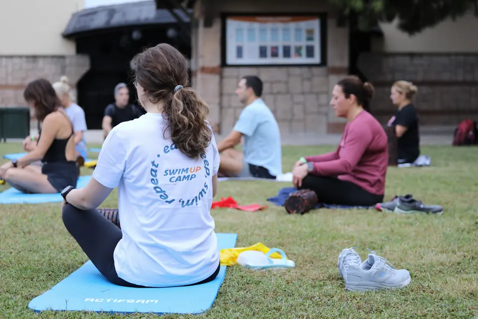 Çimde bağdaş kuran grup, arkadan görünen bir katılımcı Swim Up Camp tişörtüyle matta oturuyor.