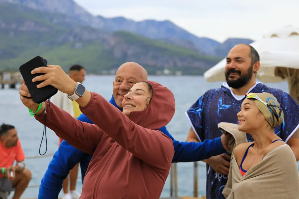 Havlu ve pançolara sarınmış bir grup, biri çekim yaparken denizin önünde poz veriyor.