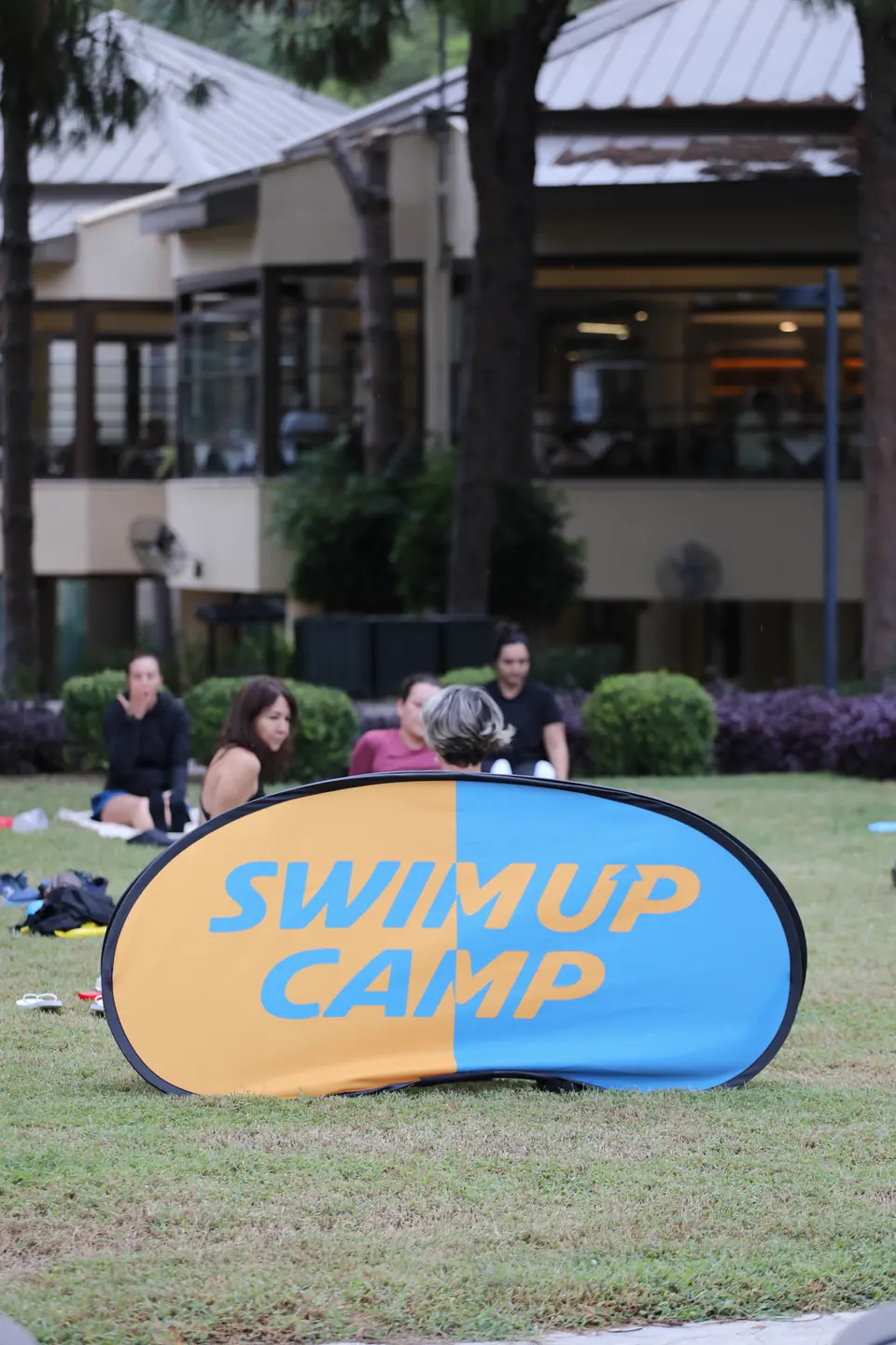 Çimdeki yoga matlarının arkasında Swim Up Camp tabelası, uzakta otelin teras katı.
