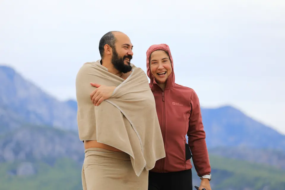 Havluya sarınmış iki kişi omuz omuza gülümsüyor; arkada dağlar ve deniz.