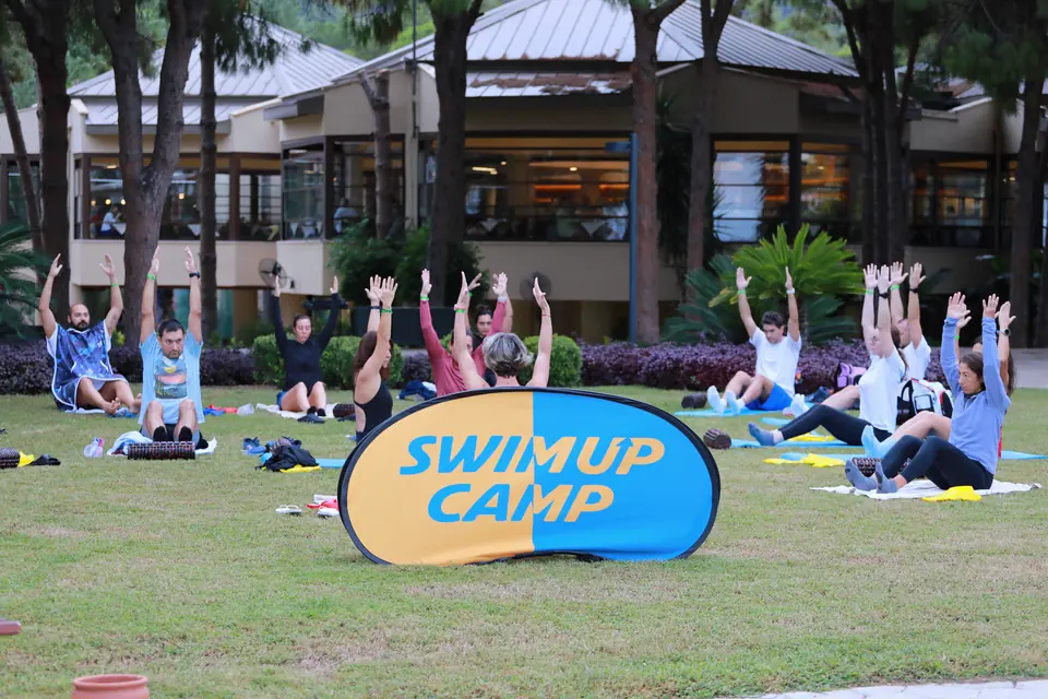 Çimde bir grup, kollar yukarıda ortak esneme yapıyor; ortada Swim Up Camp tabelası var.