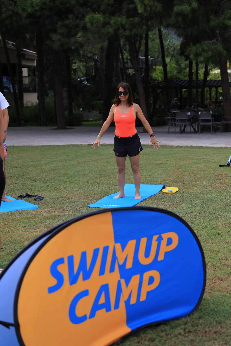 Çimde mat üzerinde tek başına ayakta ısınan katılımcı; önde Swim Up Camp tabelası.