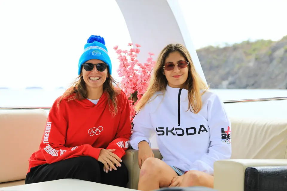 Teknenin güvertesinde iki kişi yan yana oturuyor; biri mavi bere ve kırmızı sweatshirt giyiyor.