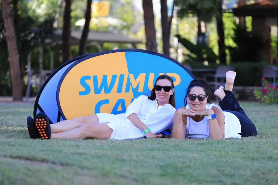 İki katılımcı çimde Swim Up Camp tabelasının önünde yan yana uzanmış, gülümsüyor.