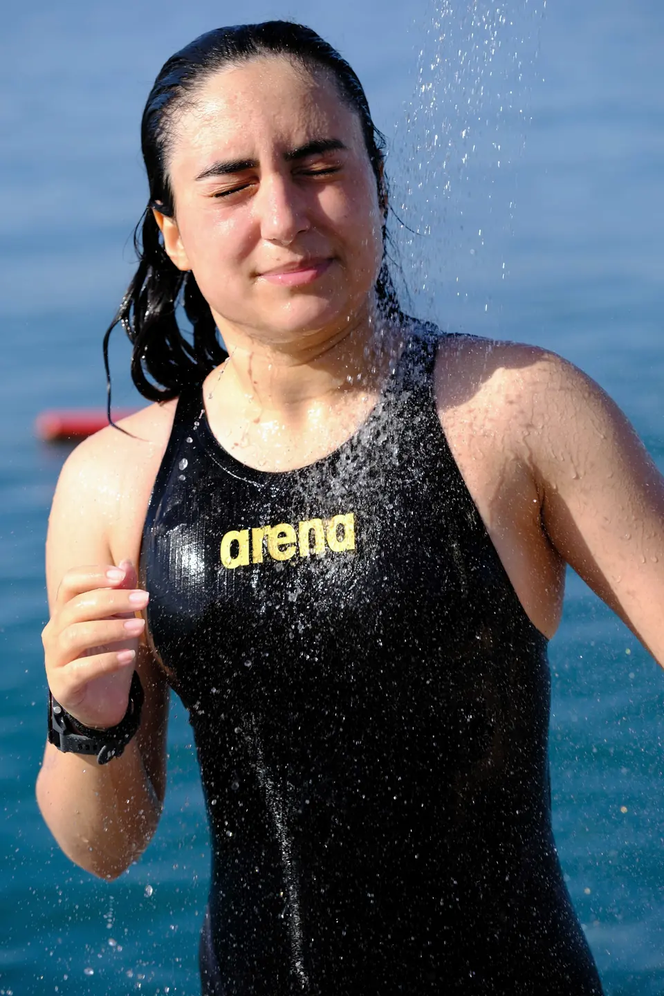Islak mayolu yüzücü plaj duşundan sonra gözleri kısık duruyor; arkada deniz.