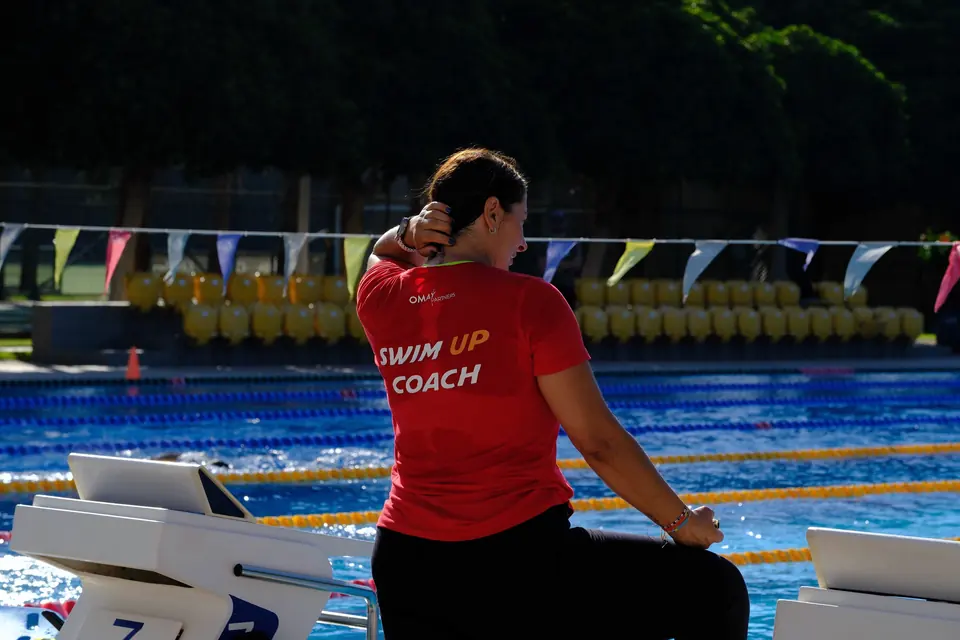 Kırmızı Swim Up Coach tişörtlü antrenör, start kürsüsüne oturmuş havuzu izliyor.