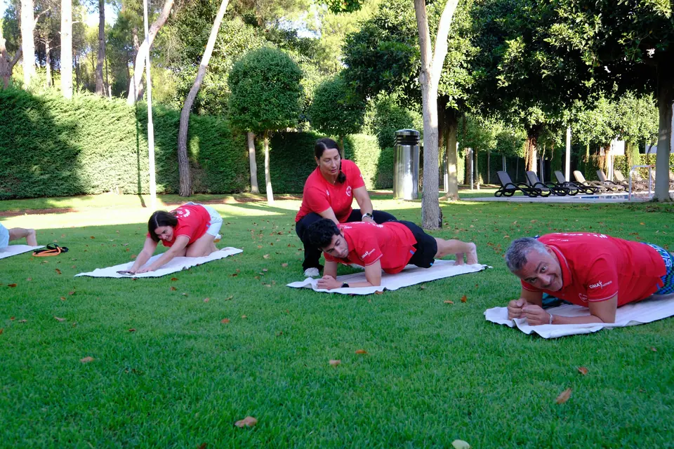 Çimde havlu üzerinde plank ve esneme yapan katılımcılar, bir antrenör düzeltme yapıyor.