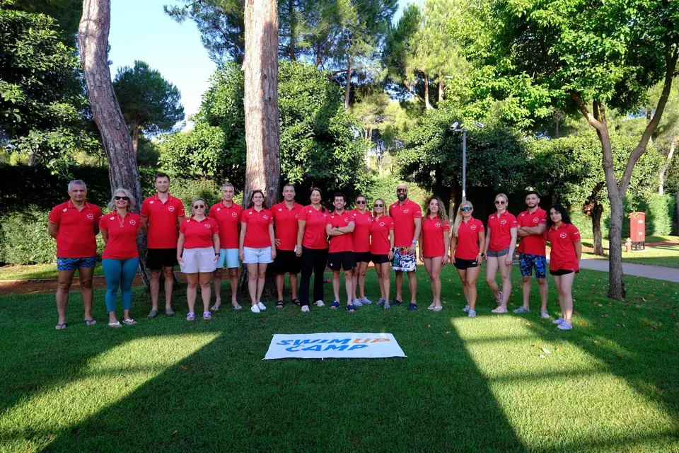 Kırmızı tişörtlü ekip çimde Swim Up Camp tabelasının arkasında dizilmiş poz veriyor.