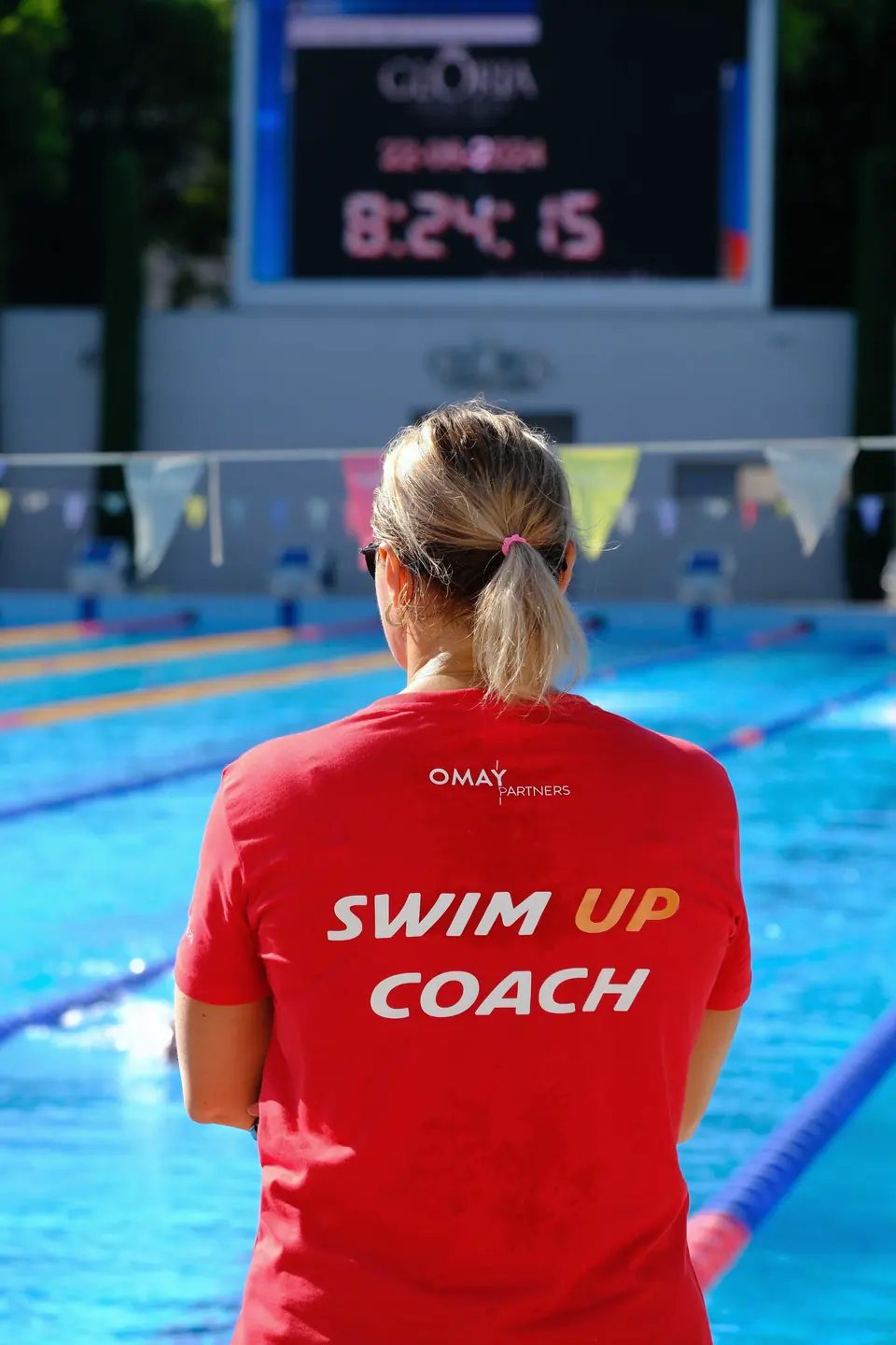 Kırmızı Swim Up Coach tişörtlü antrenör havuz kenarında arkadan, zaman panosuna bakıyor.