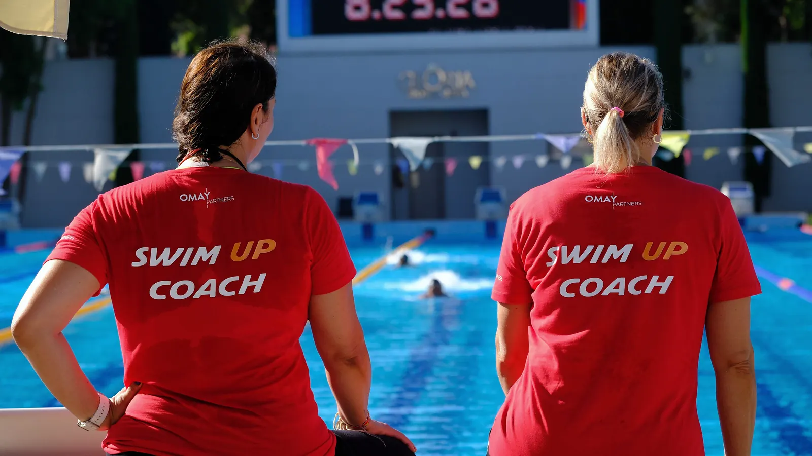 Haziran 2024, Gloria Sports Arena: SWIM UP COACH tişörtlü iki antrenör, havuz başında kulvardaki yüzücüleri izliyor.